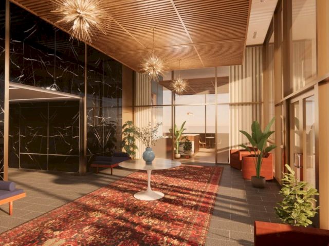 A modern hotel-like lobby with a red patterned rug, plants, seating, and sparkling chandeliers under warm ceilings.